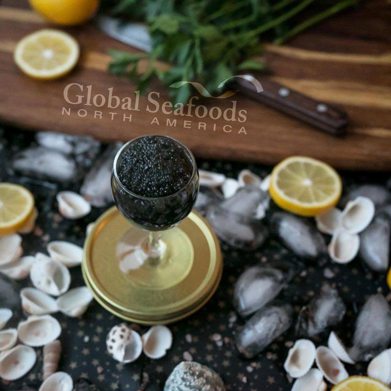 Angled overhead view of Sevruga caviar in a stemmed glass on a gold tin, with ice, shells and a cutting board