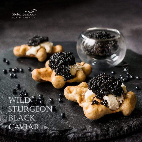Hackleback caviar on three pastries with cream on a slate board, with a glass bowl of caviar behind them
