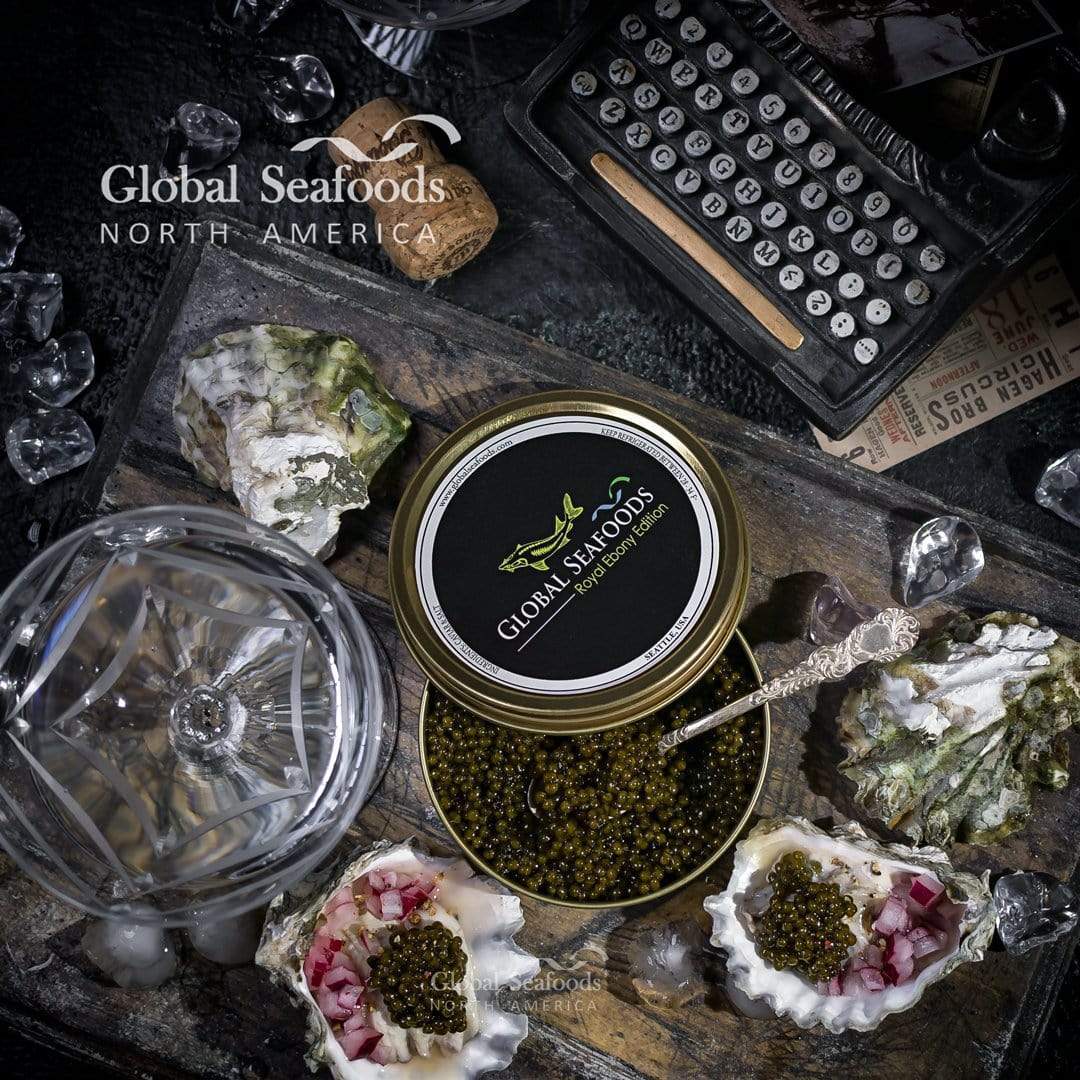 Kaluga caviar tin with a spoon on a wooden board beside oysters topped with caviar and diced red onion