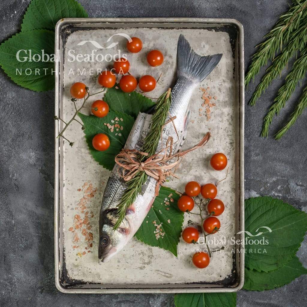 Whole branzino in a metal tray with tomatoes, herbs and green leaves.