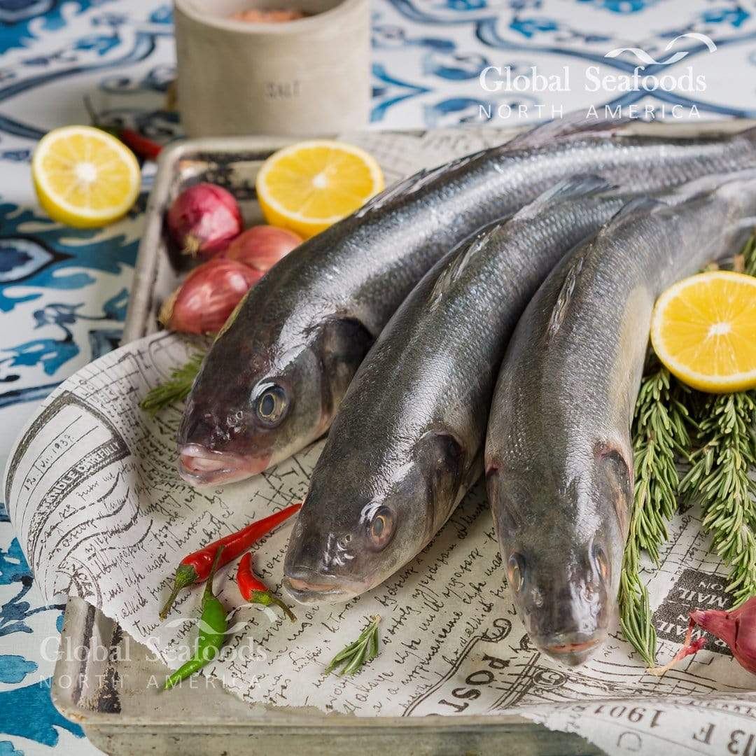 Three whole branzino on paper in a tray with lemon and shallots.