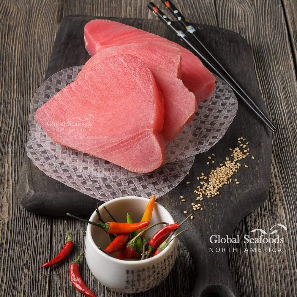 Three fresh Ahi tuna steaks arranged on a brown cutting board over a brown table, with chopsticks on the side and a small cup containing chili peppers for a bold, Asian-style seafood presentation