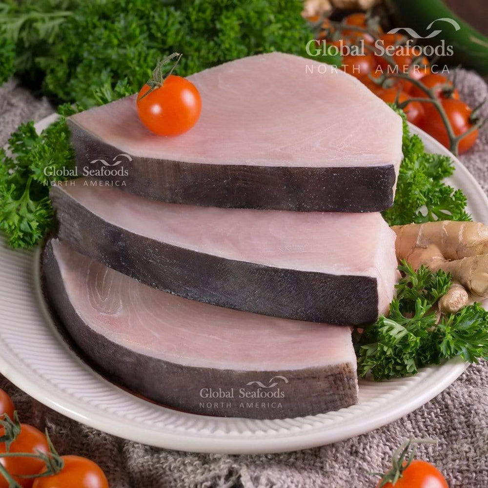 three swordfish steaks turned skin-side up on a white plate, surrounded by cherry tomatoes with leafy greens in the background, styled for a fresh and colorful seafood presentation