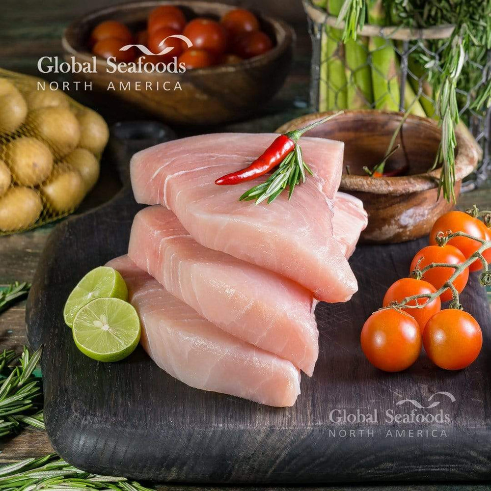 Three fresh swordfish steaks on a black stone surface, surrounded by lime slices, cherry tomatoes, small potatoes, and asparagus for a colorful and wholesome seafood display
