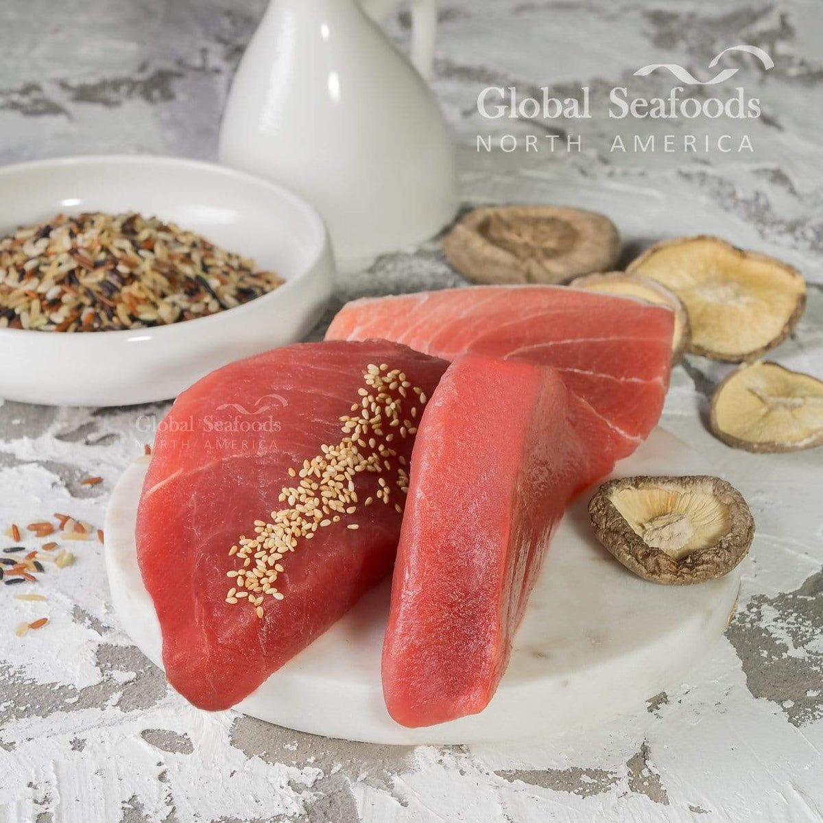 Two raw yellowfin tuna steaks on a white board, one topped with sesame seeds, beside dried mushrooms and rice.