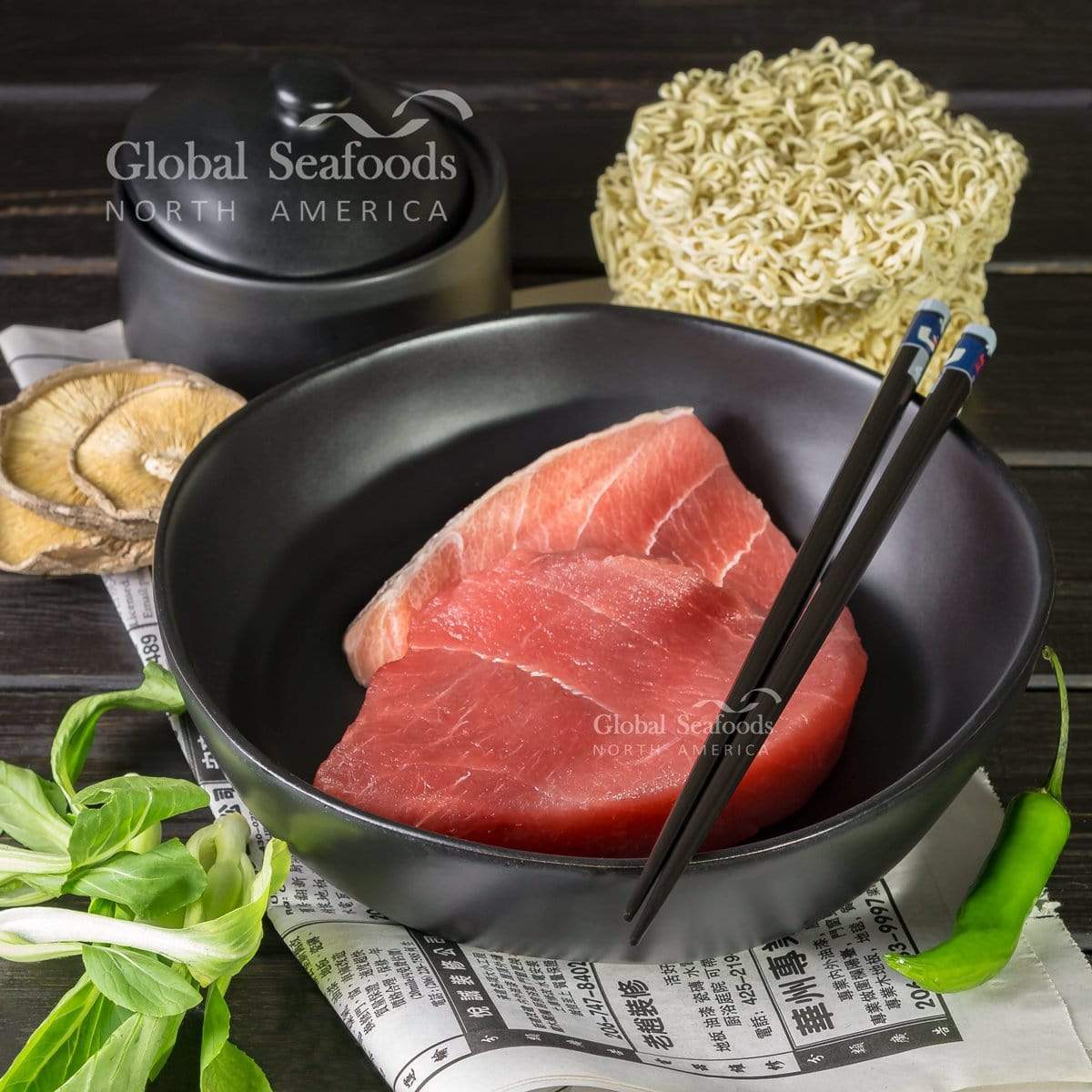 Raw yellowfin tuna steak in a black bowl with chopsticks, beside dried noodles and mushrooms.