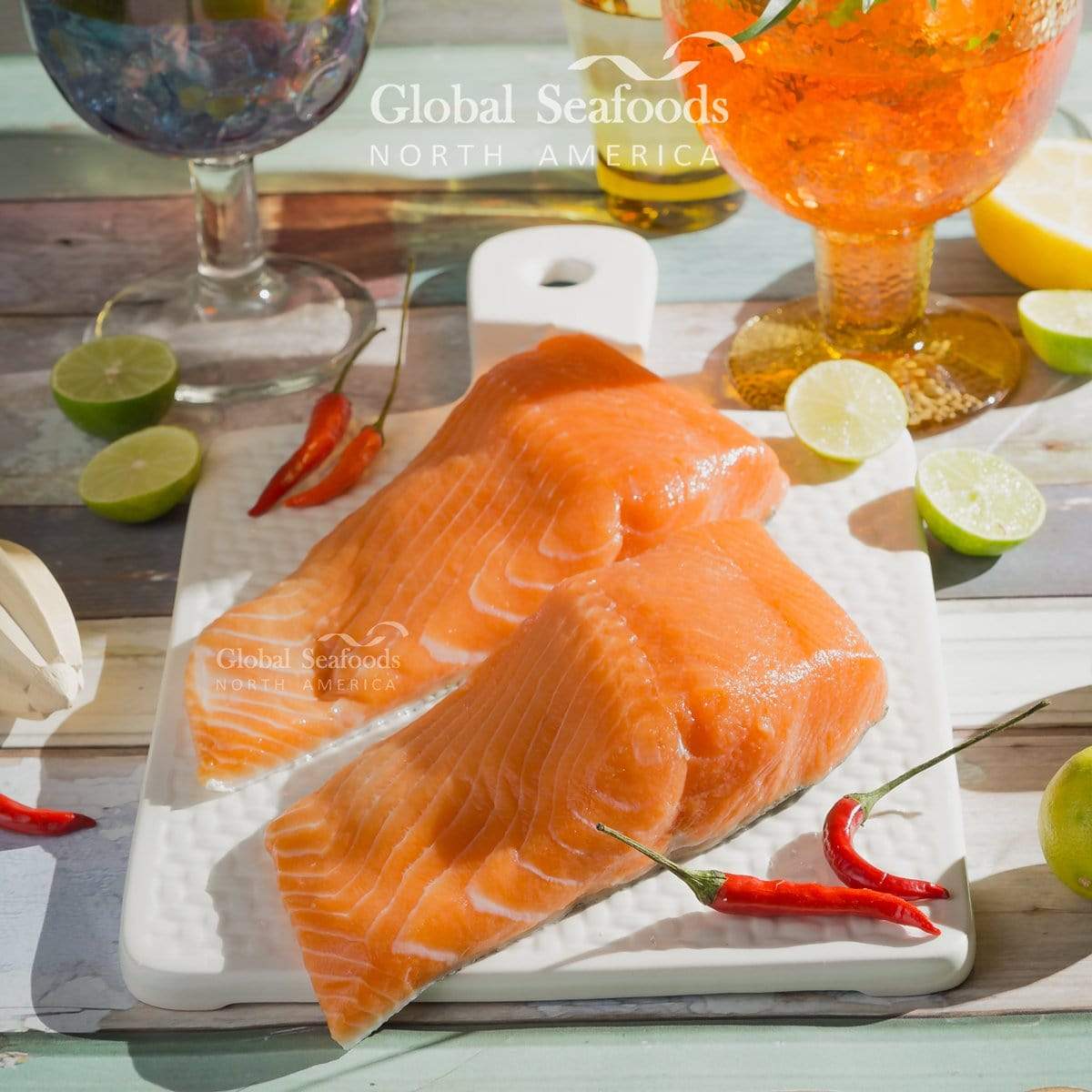 Two raw skin-on king salmon fillets on a white cutting board with red chilies, lime and lemon.
