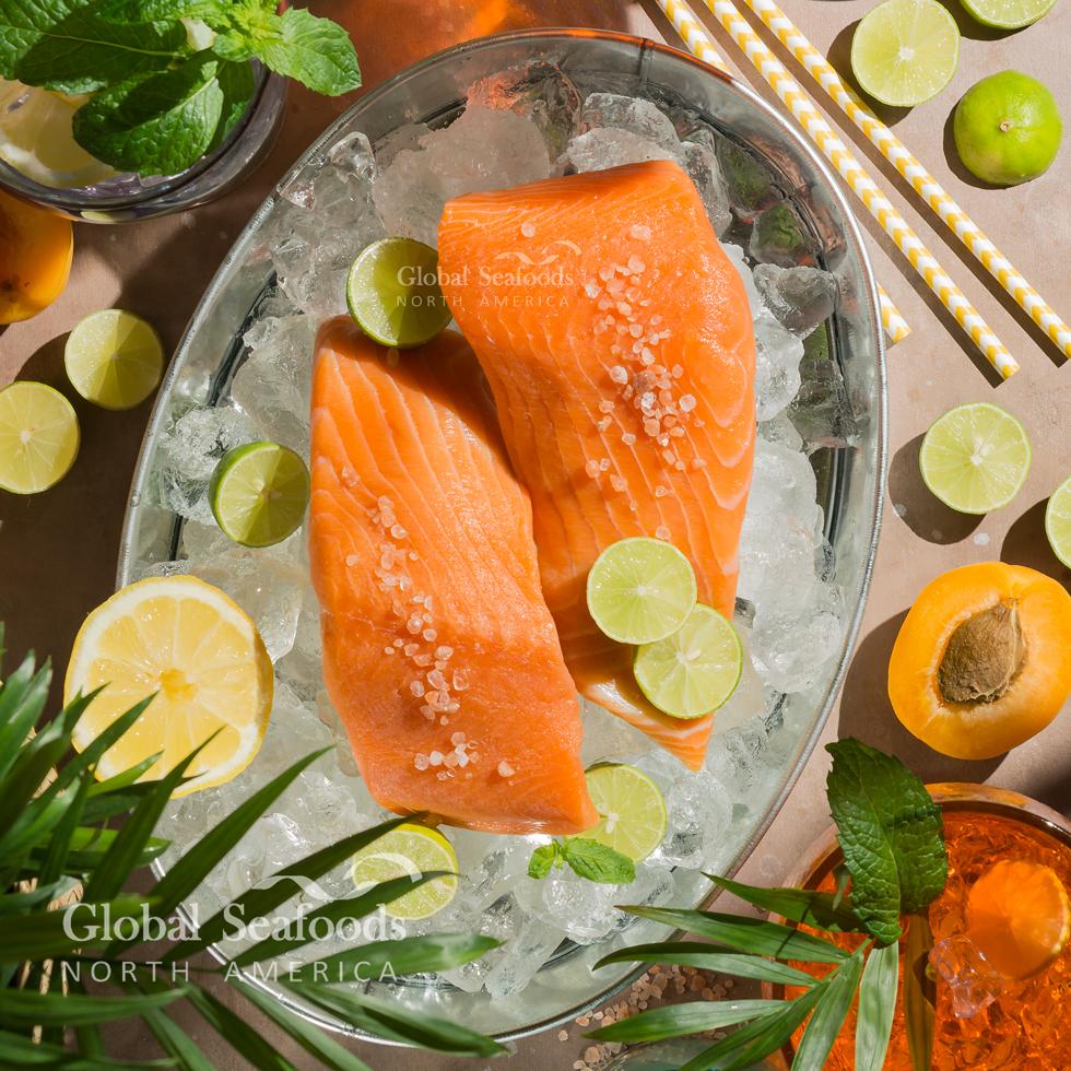 Two raw king salmon fillets on crushed ice in a metal platter with lemon and lime slices.