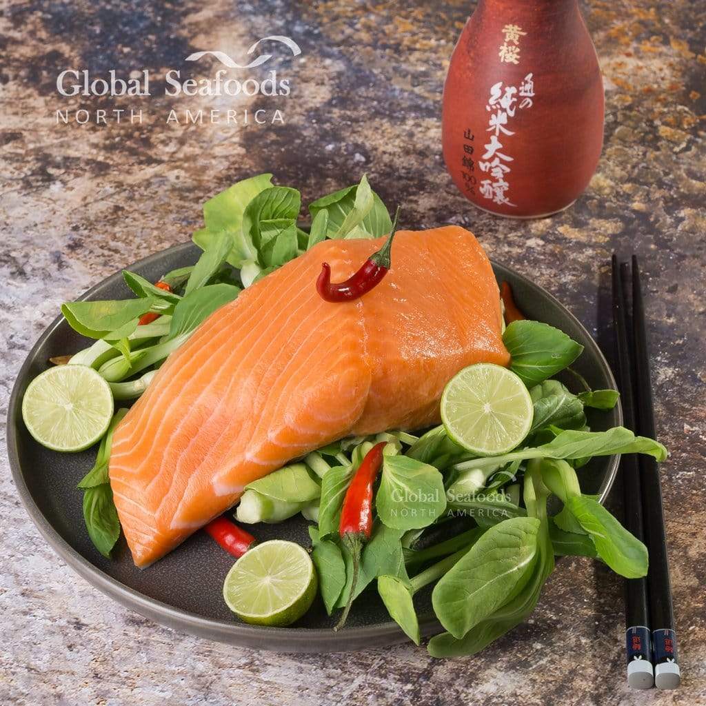 Raw king salmon fillet on leafy greens with lime and red chilies, served on a dark plate beside chopsticks.