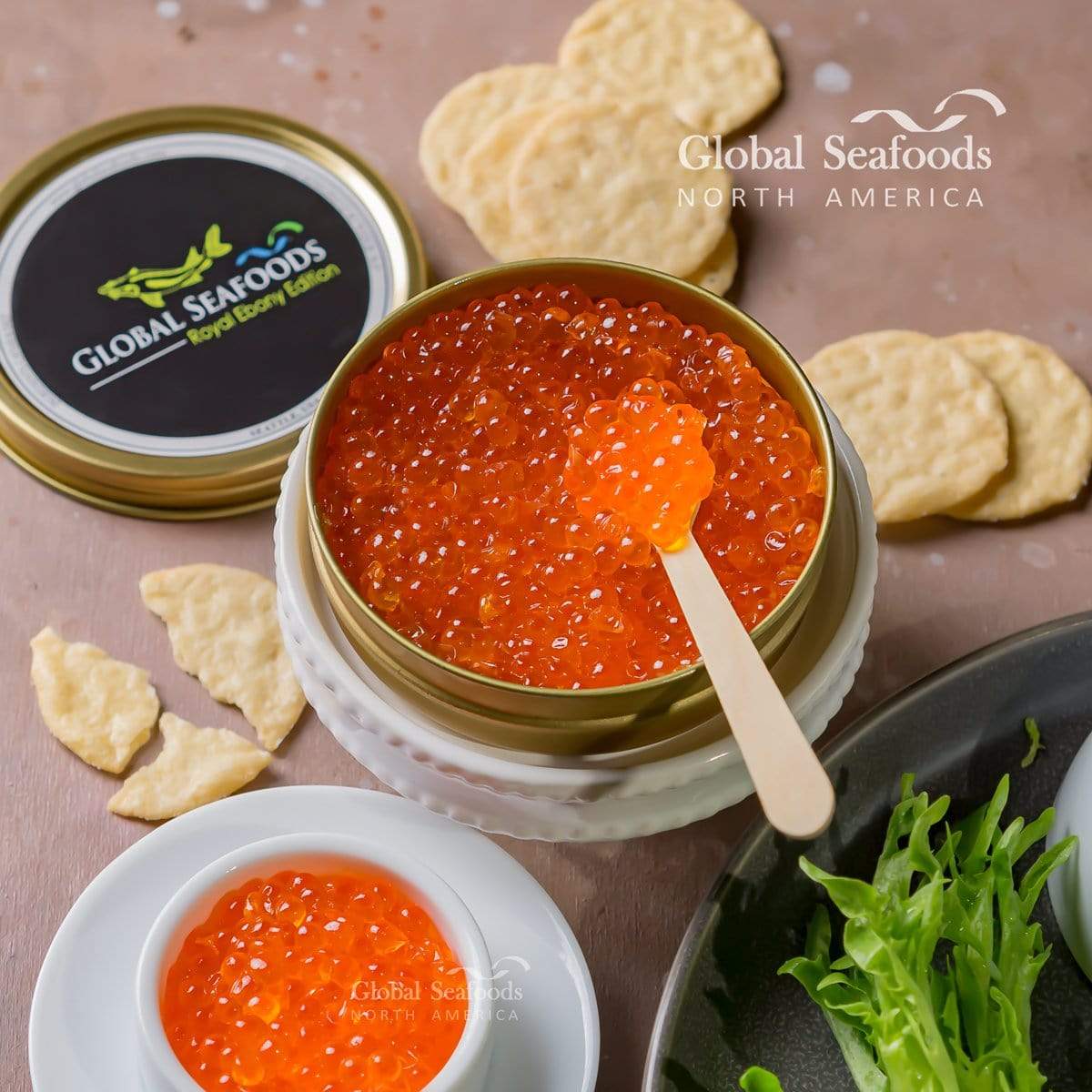 Open gold tin of orange trout roe with a wooden spoon, beside crackers and a small white bowl of roe