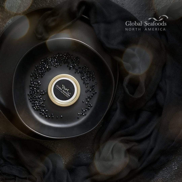 Overhead view of a closed Beluga hybrid caviar tin surrounded by scattered caviar pearls on a black plate