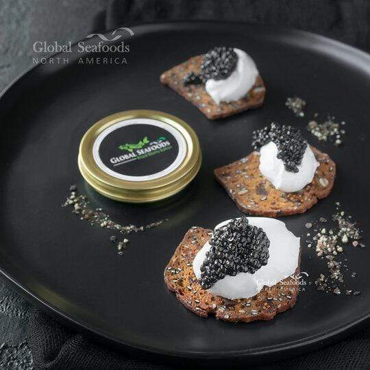 Beluga hybrid caviar served on three crackers with sour cream, arranged on a black plate alongside an open caviar tin for a luxurious appetizer presentation