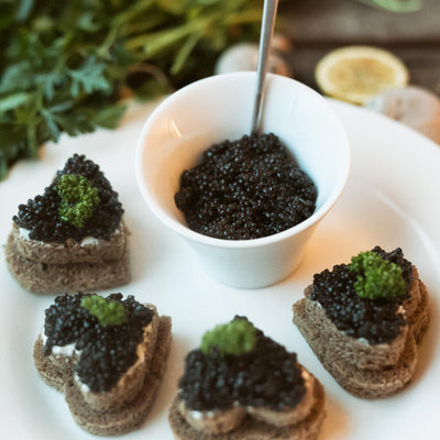 Premium California White Sturgeon Caviar served in a white cup with a spoon, accompanied by four heart-shaped brown bread toasts, two of which are topped with caviar for an elegant and romantic presentation