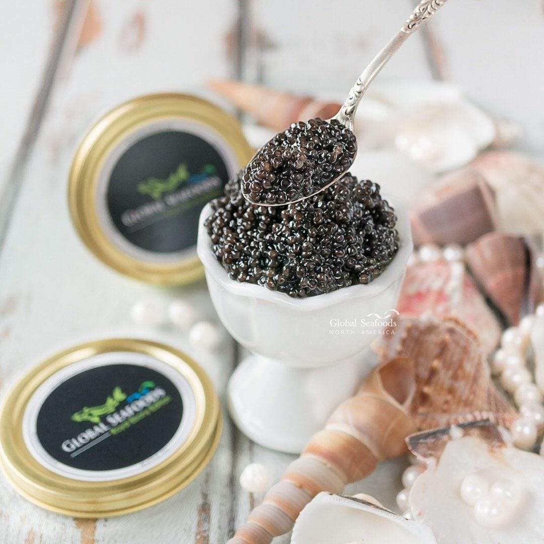 Spoonful of Siberian sturgeon caviar above a white egg cup, with a closed tin and seashells
