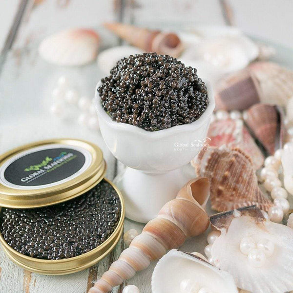 Siberian sturgeon caviar in a white egg cup beside an open tin, with seashells on a white wooden surface