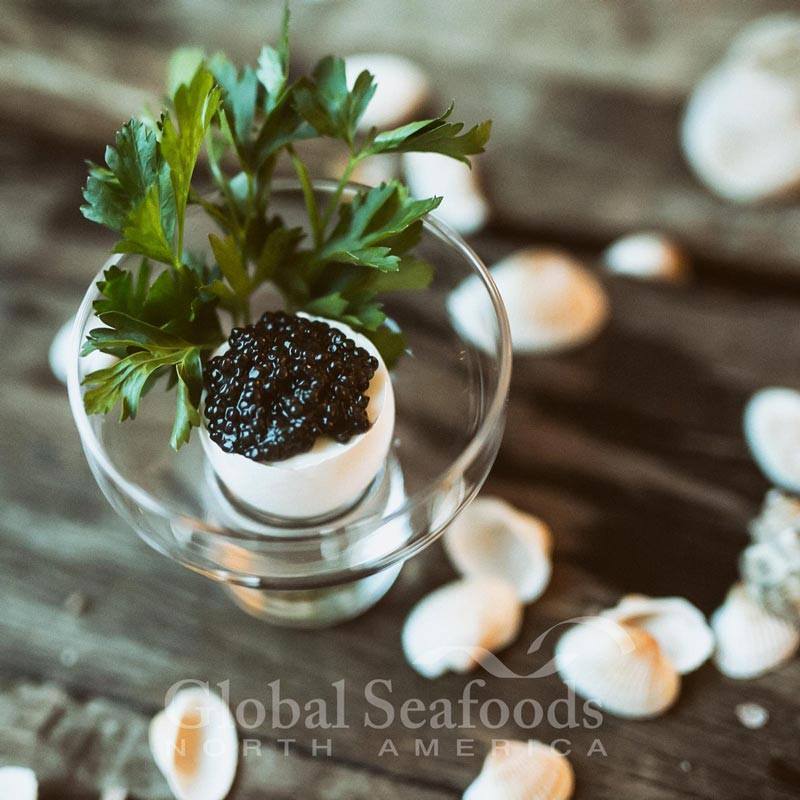 White sturgeon caviar served in an eggshell in a glass dish with parsley and seashells on a wooden table