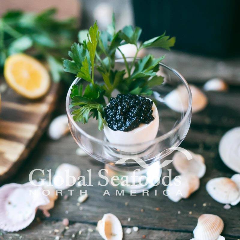 White sturgeon caviar in an eggshell with parsley, displayed in a glass dish beside seashells and lemon