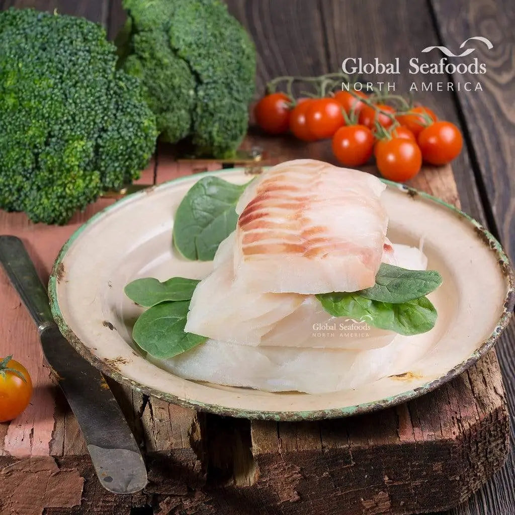 Pacific cod fillet portions on a plate with green leaves, with broccoli and tomatoes behind.