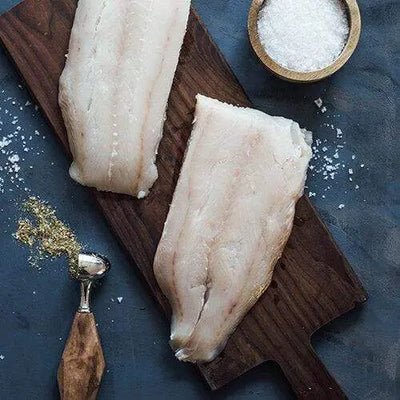 Two Lingcod fillets placed on a wooden board with a small cup of white salt beside them, styled for a clean and rustic seafood preparation