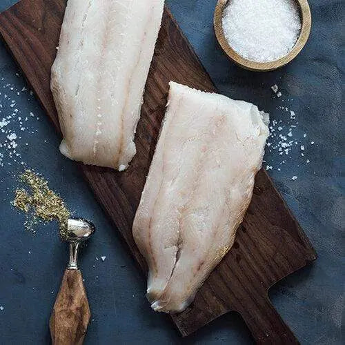 Two raw lingcod fillets on a wooden board beside a bowl of coarse salt