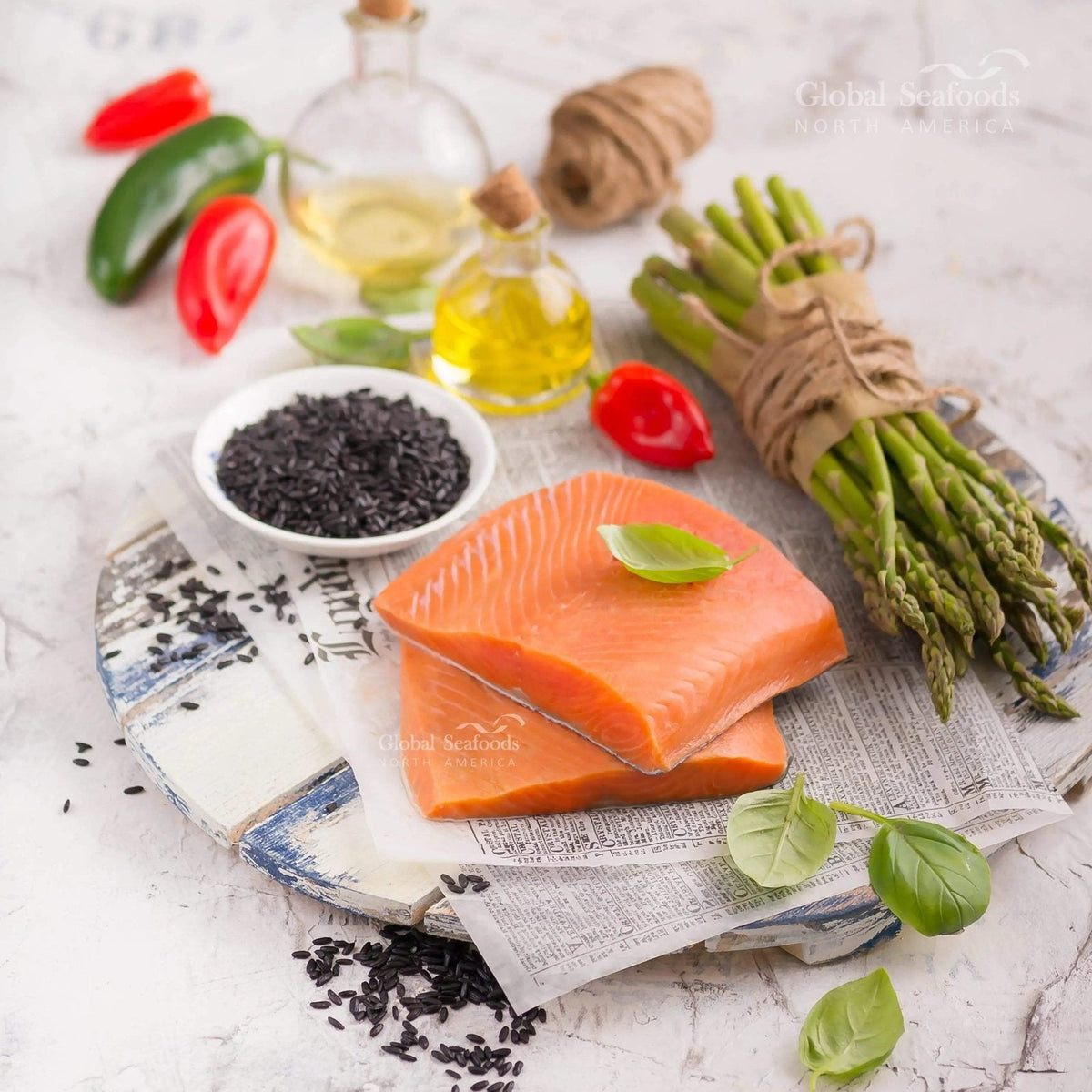 Two raw skin-on sockeye salmon portions on printed paper over a wooden board, beside asparagus, black rice and basil.