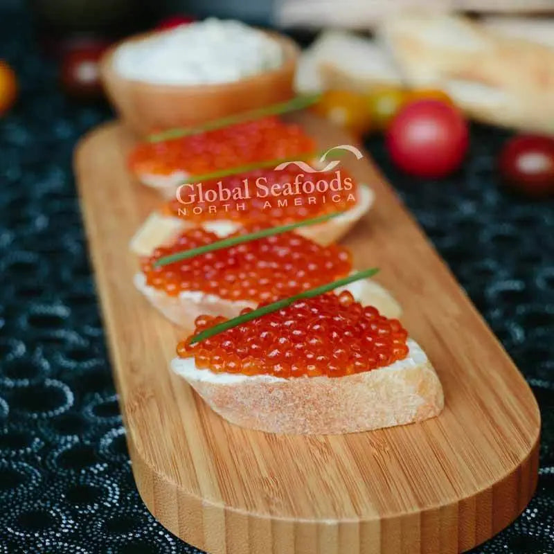 Four baguette slices topped with pink salmon caviar and chives on a wooden serving board
