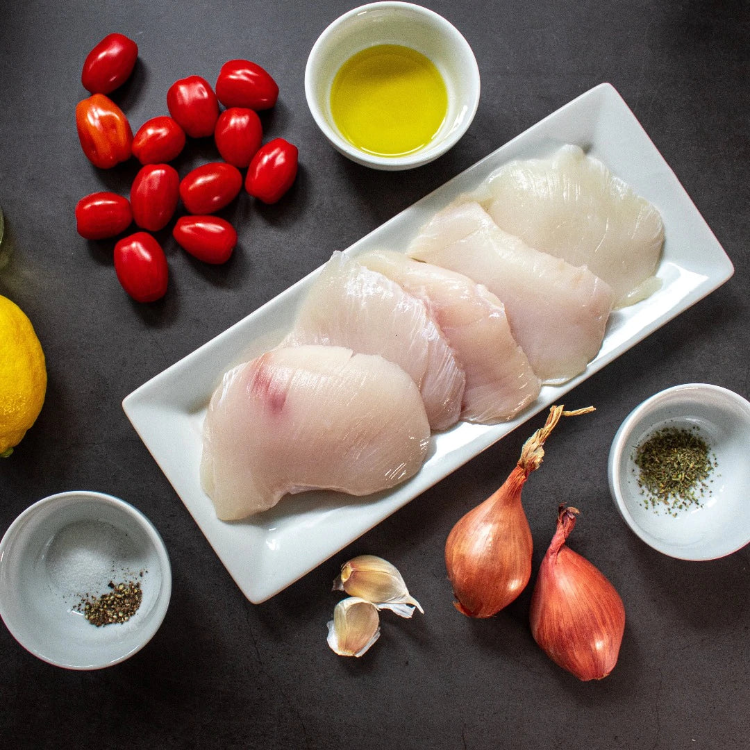 Raw halibut cheeks on a rectangular white plate with tomatoes, shallots and garlic.